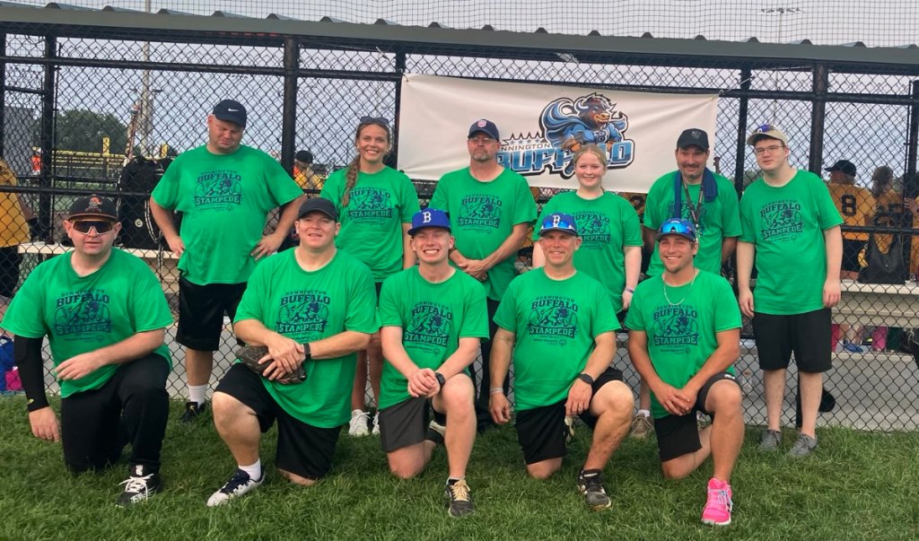 A group of ten people wearing green Buffalo Stampede t-shirts pose for a photo in front of a sports field and a team banner.