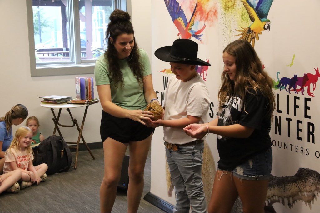 A presenter shows an animal to a group of children during an educational event, with three children actively participating and observing.