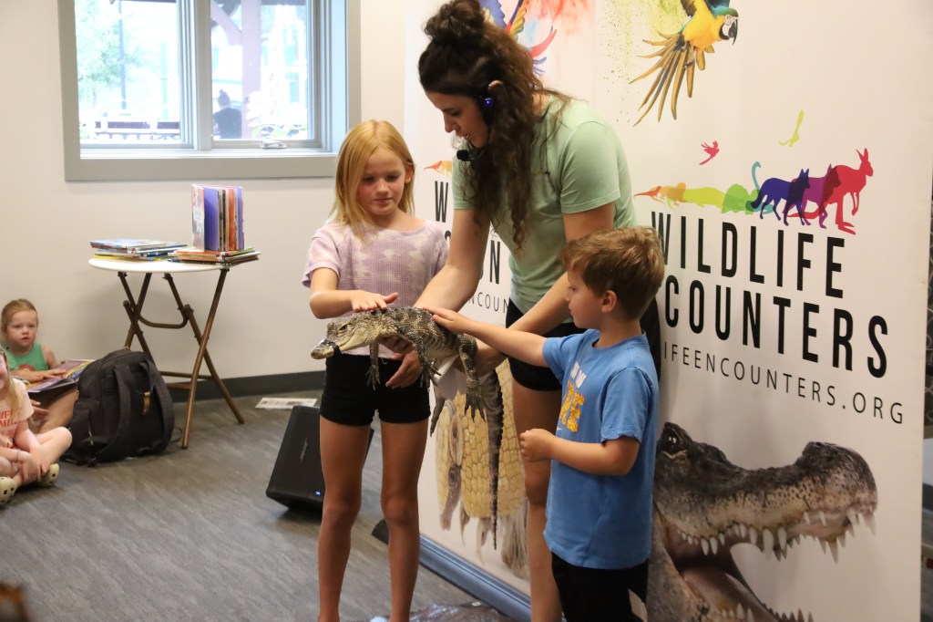 A child and a young girl interact with a small alligator being held by a woman at a wildlife education event, with a banner in the background displaying 'Wildlife Encounters.'