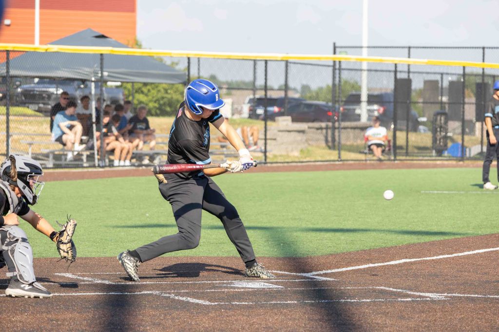 A young baseball player in a blue helmet swings a bat at a pitch on a baseball field, with a catcher and spectators in the background.