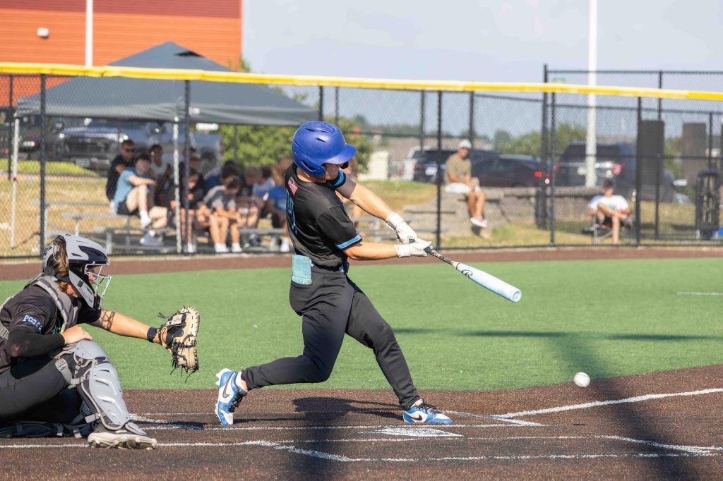 A baseball player in a blue helmet swings a bat at a pitch while a catcher in gray gear prepares to catch the ball. Spectators are seated in the background.