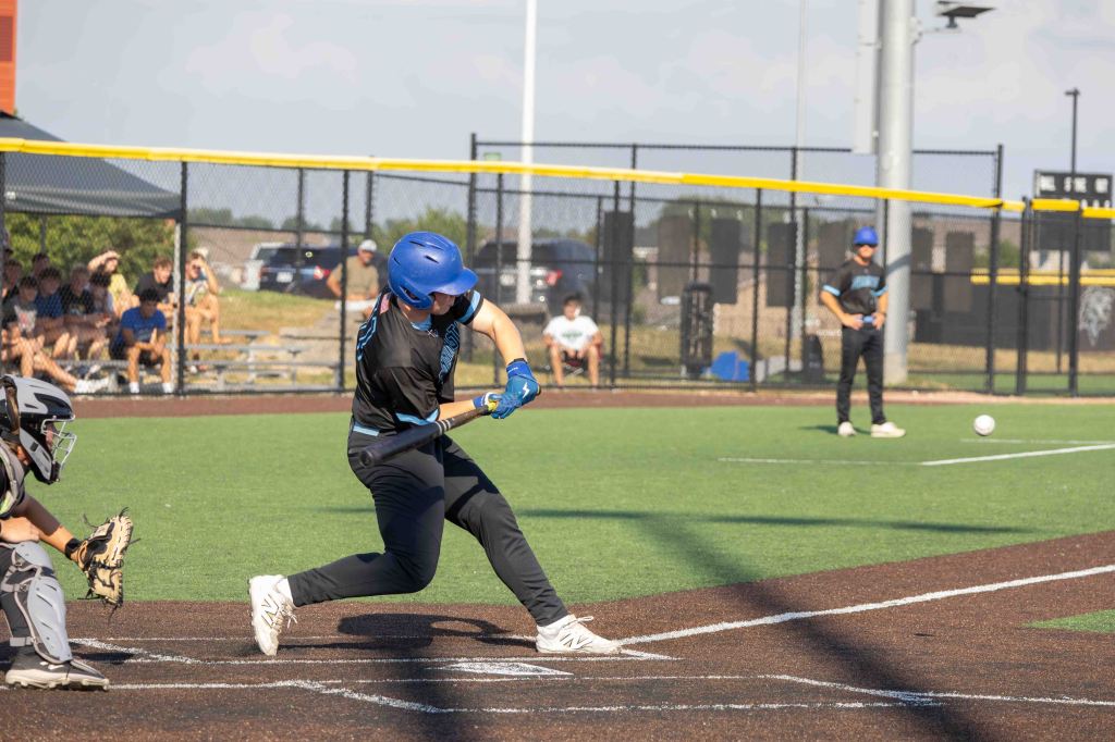A baseball player swings at a pitch during a game, with a catcher preparing to receive the ball. Spectators are seated in the background.