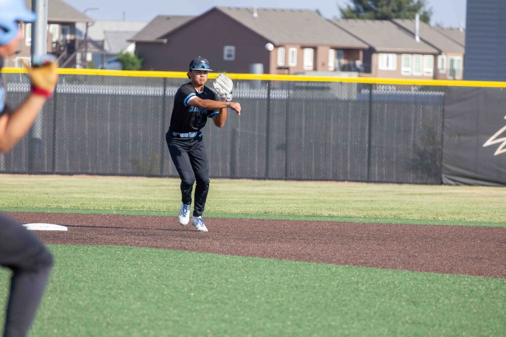 A baseball player in a black uniform prepares to throw a baseball while standing on a field. In the foreground, another player is visible, also in a uniform, as they focus on the action.