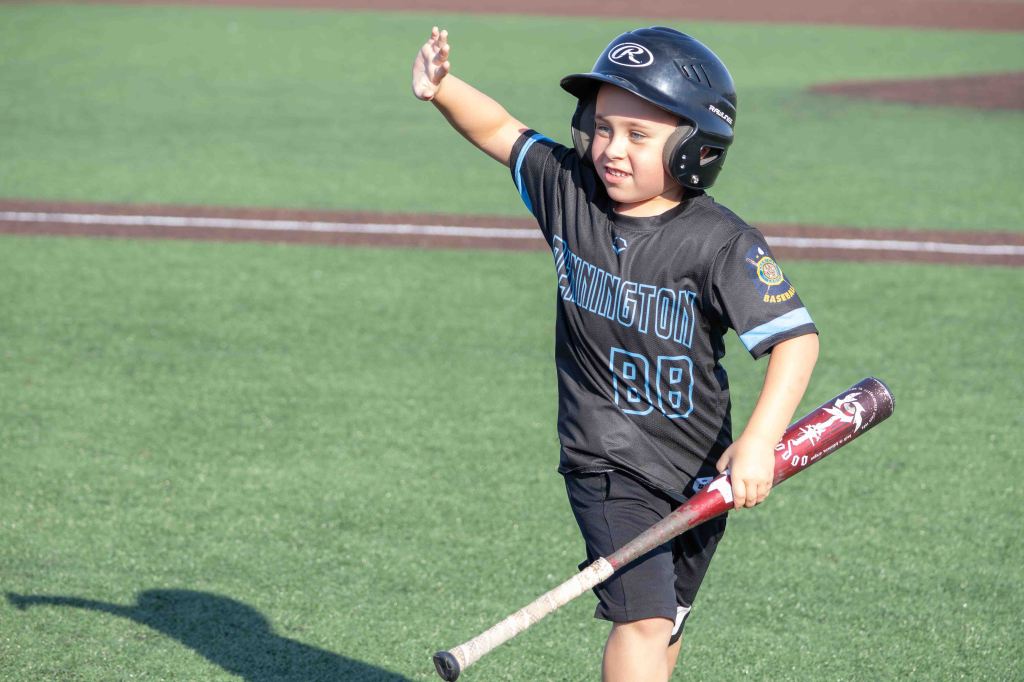 A young boy in a black baseball jersey with 'Runnington' printed on it and the number 88, wearing a batting helmet, joyfully waves while holding a baseball bat on a grassy field.