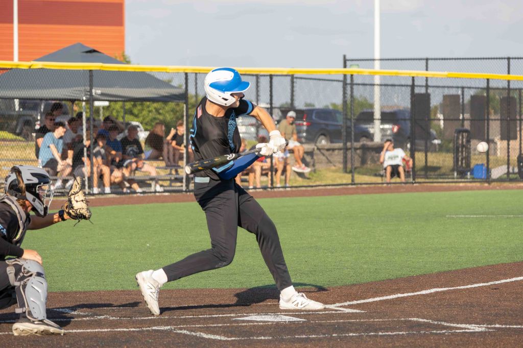 A young baseball player in a blue helmet is swinging a bat during a game, with a catcher in protective gear behind him and spectators in the background.