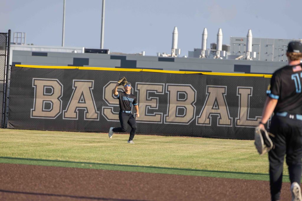 A baseball player running toward the outfield fence, wearing a black uniform with blue accents, preparing to catch a fly ball. The fence features the word 'BASEBALL' prominently displayed.