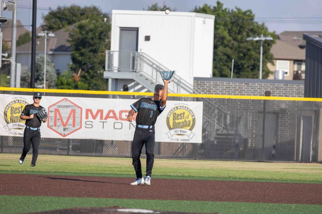 A baseball player in a black uniform catches a fly ball while standing on a grassy field. Another player is in the background, ready for action.