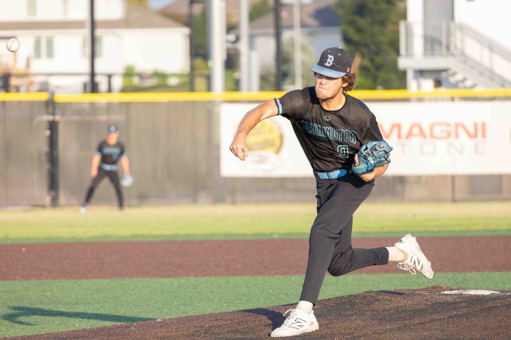 A baseball player in a black jersey with 'Bennington' written on it, throwing a pitch on a baseball field, with a second player in the background.