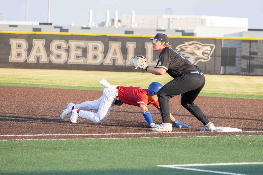 A baseball player in a red jersey dives towards first base while an opposing player in a black jersey prepares to tag him out.