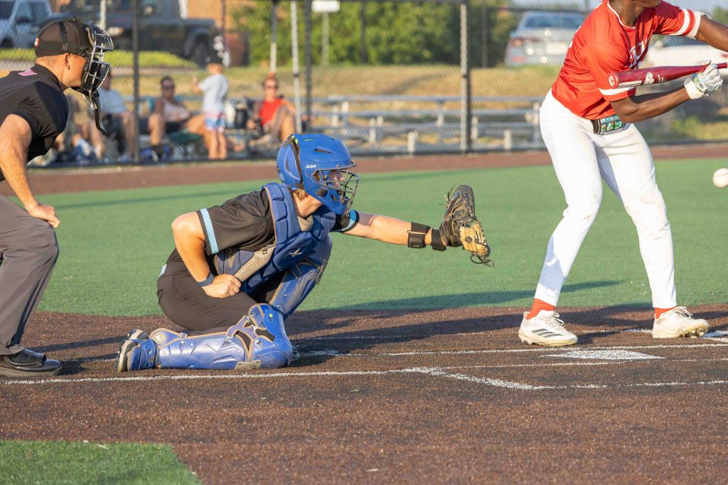 A baseball catcher in blue gear attempts to catch a pitch from a batter in red, while an umpire watches from behind.