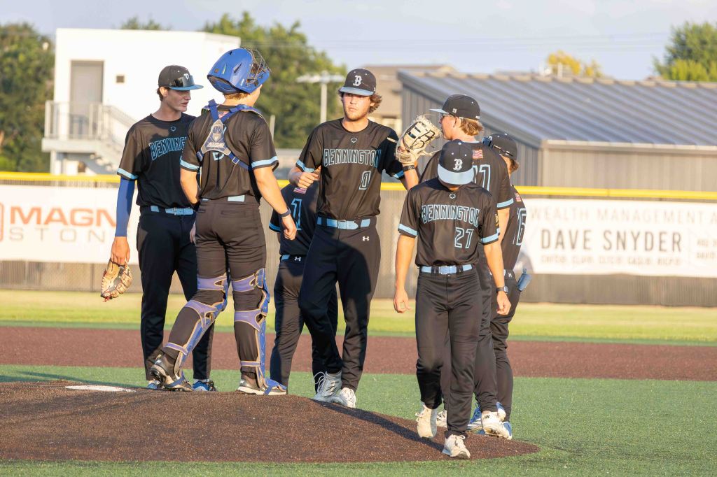 A group of young baseball players in black uniforms with blue accents gathered on the pitcher's mound for a team meeting.