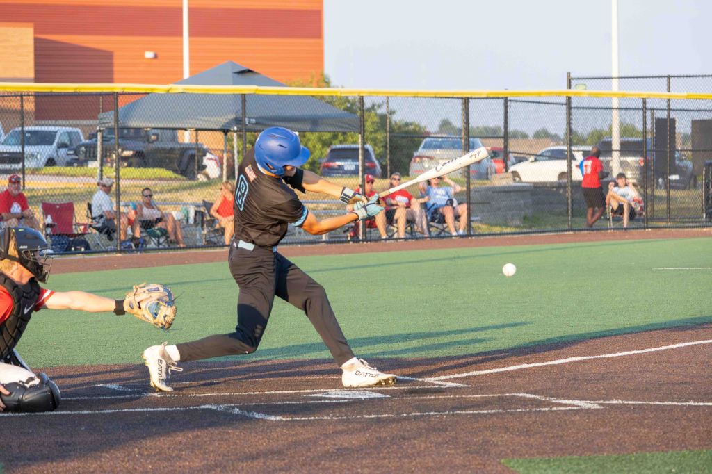 A young baseball player in a blue helmet swings at a pitched ball, while a catcher in black gear prepares to receive it. The scene is set on a baseball field with spectators watching in the background.