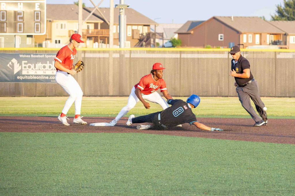 A baseball play at second base, with a player in black sliding into the base while a player in red attempts to tag him. An umpire observes the play in the background.