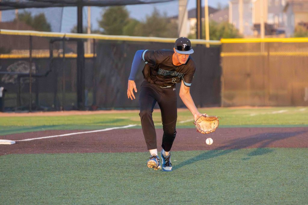A baseball player in a black uniform is fielding a ground ball on the baseball diamond during sunset.