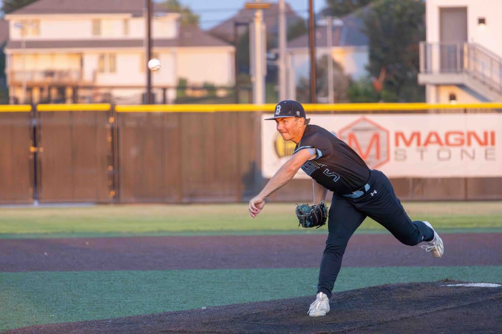 A baseball pitcher in a black uniform throwing a baseball on a field during sunset.