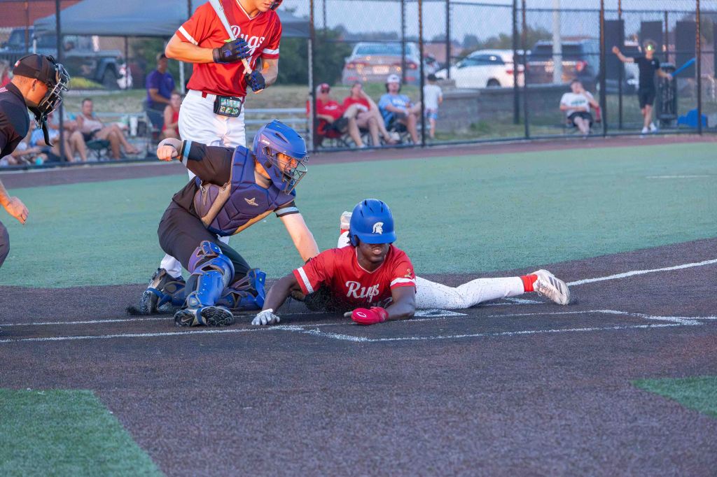 A baseball player in a red jersey slides towards home plate while a catcher in blue gear prepares to tag him. The background features spectators watching the game.