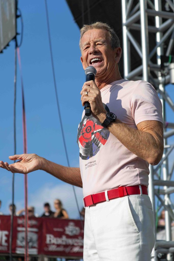 A man in a light pink shirt with a graphic design and white pants, holding a microphone and gesturing while speaking on stage at an outdoor event.