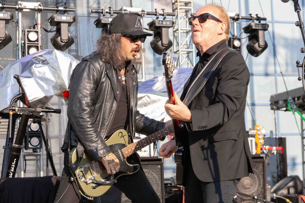 Two musicians performing on stage, one playing a guitar and the other holding an electric guitar, with sound equipment and lighting in the background.