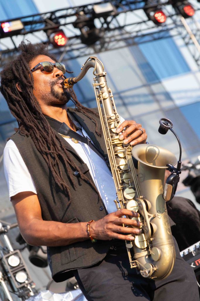 A musician playing a saxophone on stage during a performance, wearing sunglasses and a vest, with a backdrop of stage lights.