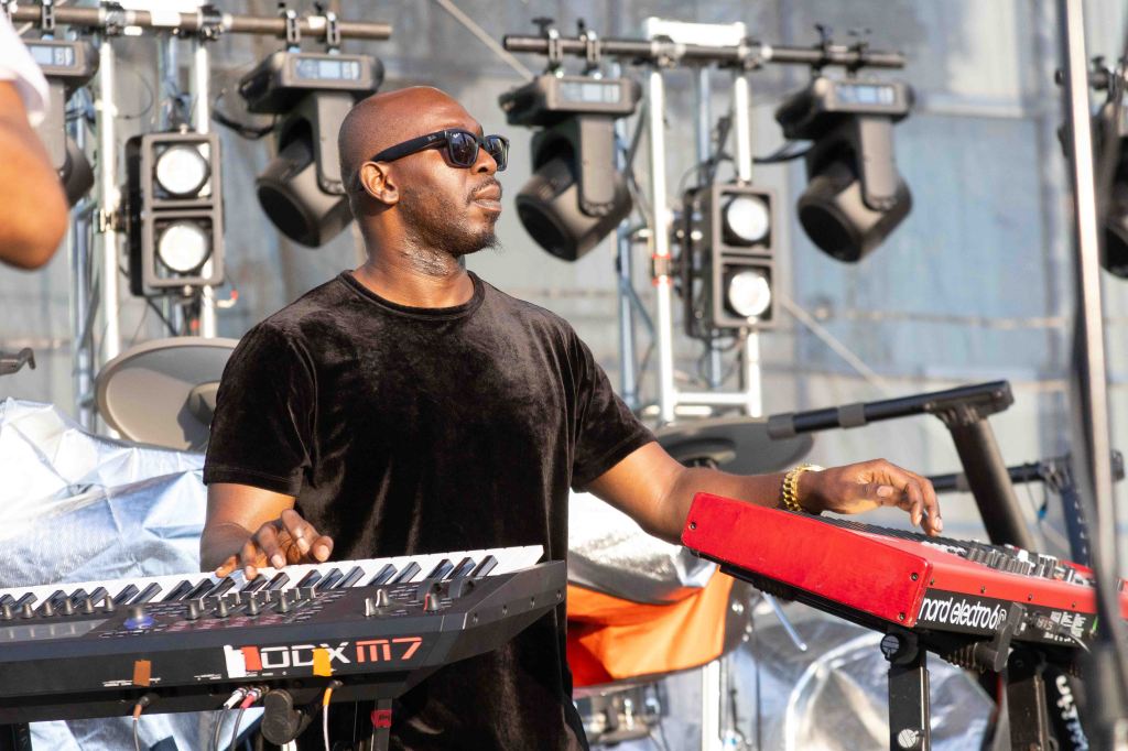 A musician playing keyboards on stage during a performance, wearing sunglasses and a black shirt, with stage lighting equipment in the background.
