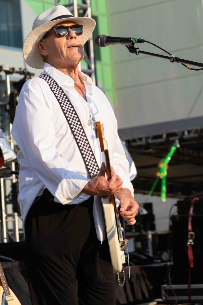 A musician performing on stage, wearing sunglasses and a white hat, holding a guitar and singing into a microphone.