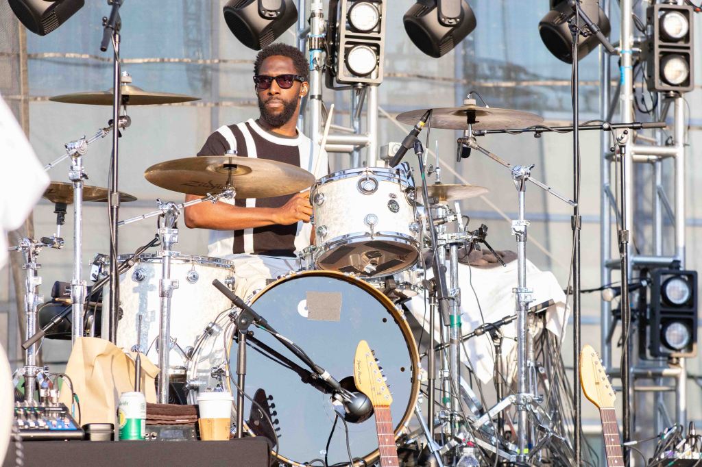 A drummer wearing sunglasses plays a white drum set on stage, surrounded by musical equipment and lighting fixtures.