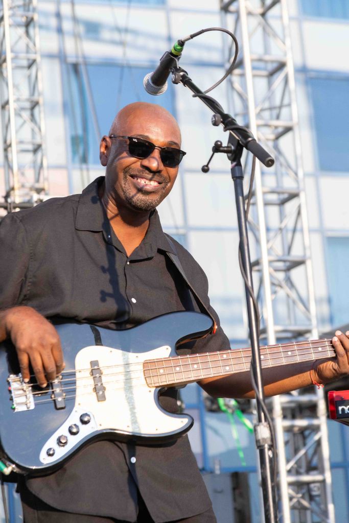 A smiling musician playing a black bass guitar on stage, wearing sunglasses and a black shirt, with microphone and sound equipment in the background.