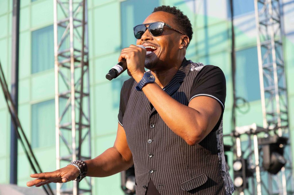 A man singing into a microphone on stage, wearing sunglasses and a stylish, dark vest, with a vibrant background of stage equipment and blurred buildings.