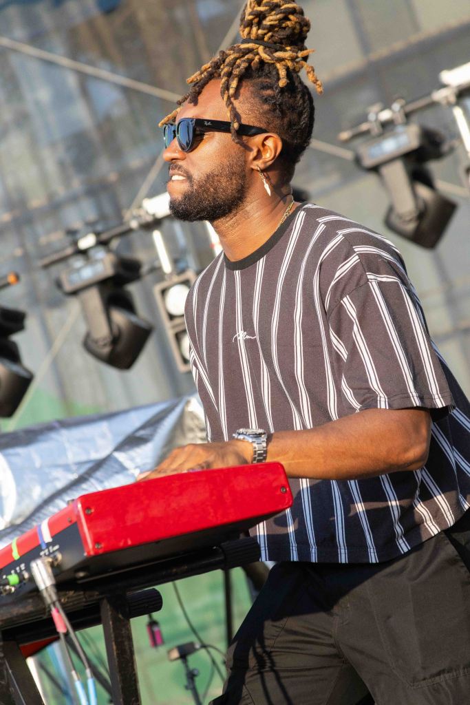 Musician playing a red keyboard on stage, wearing sunglasses and a striped shirt, with a backdrop of stage lights.