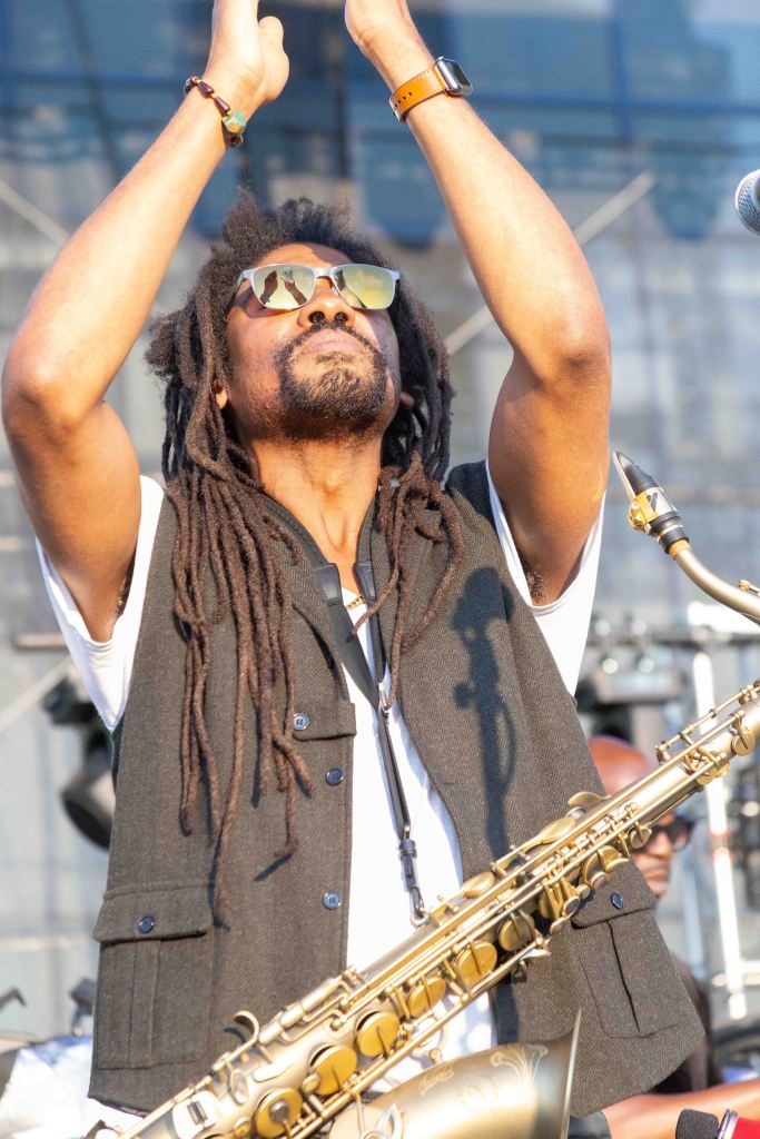 Musician with dreadlocks playing a saxophone, wearing sunglasses and a vest, raising hands in an expressive gesture on stage.