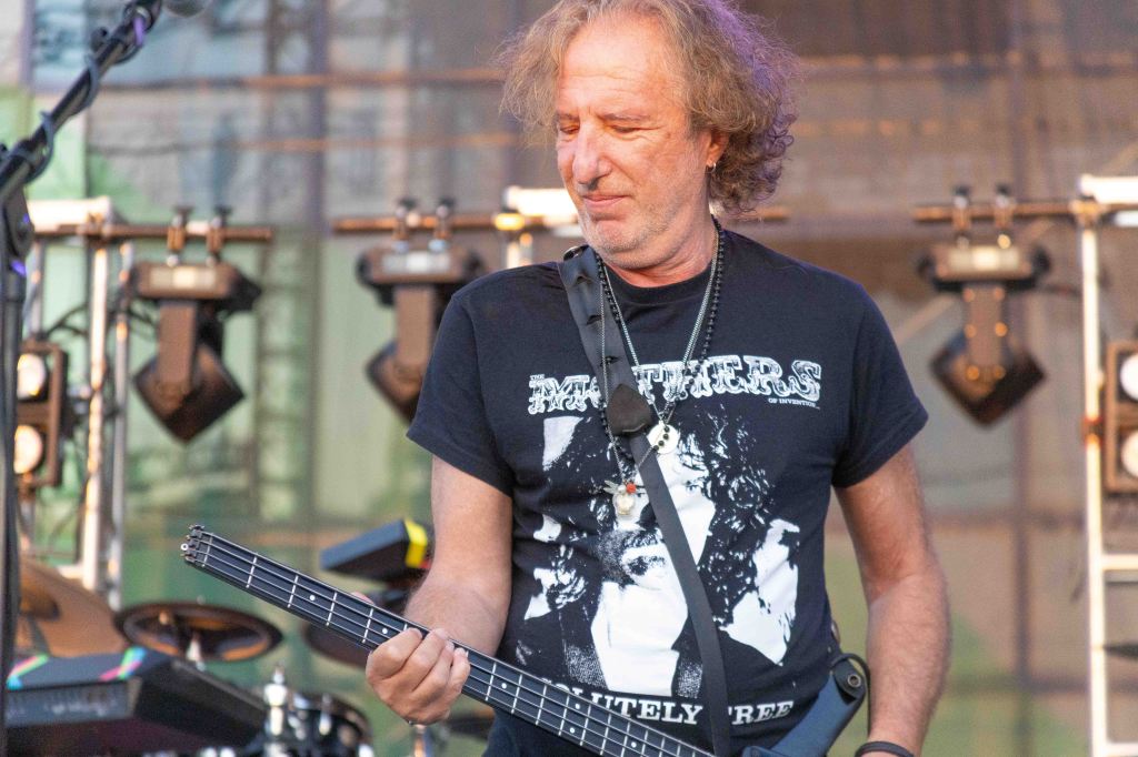 Musician playing a bass guitar on stage, wearing a band t-shirt and surrounded by stage lights and equipment.