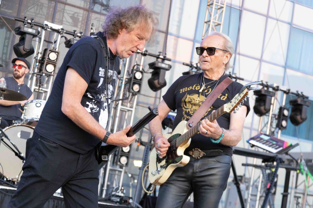 Two male musicians perform on stage with guitars, one wearing sunglasses and a black t-shirt, and the other in a graphic t-shirt. In the background, a drummer is visible.