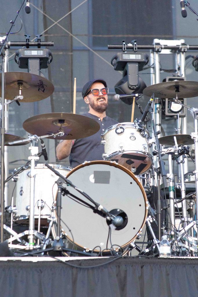A drummer performing on stage, wearing sunglasses and a cap, smiling while playing a drum set.