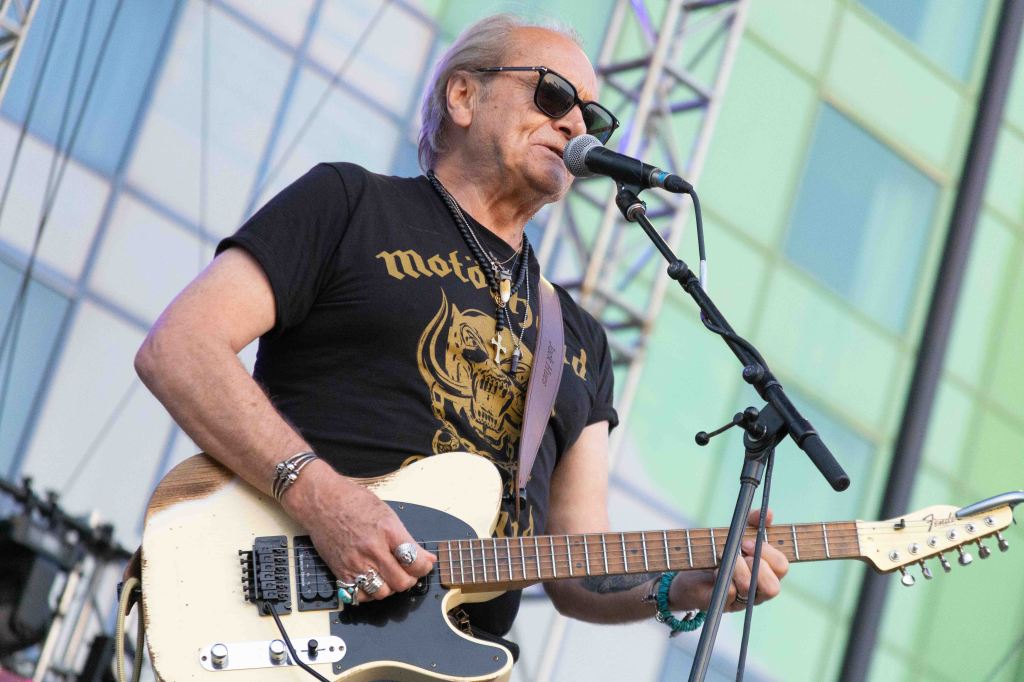 Musician performing on stage with an electric guitar, wearing sunglasses and a black t-shirt featuring a graphic design, against a colorful backdrop.
