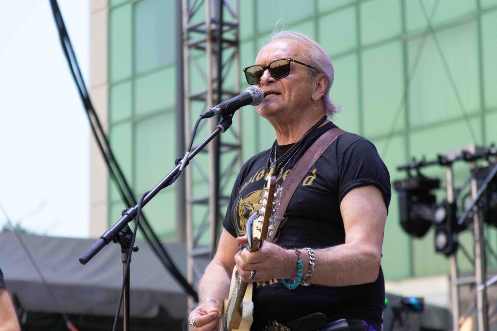 A musician with sunglasses performing on stage, holding a guitar and singing into a microphone, with a green building and lighting equipment in the background.