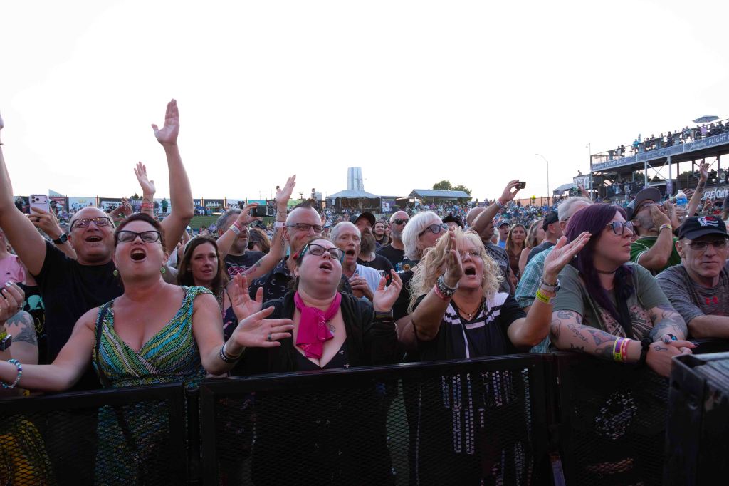 A diverse crowd of enthusiastic concertgoers singing and cheering at a live music event, with hands raised and expressions of excitement.