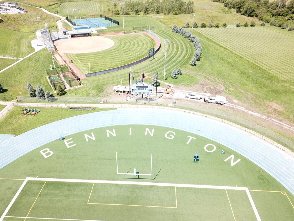 Aerial view of a sports complex featuring a running track with 'BENNINGTON' written on it, a baseball field, and various green spaces. Several trees and a parking area are visible in the background.