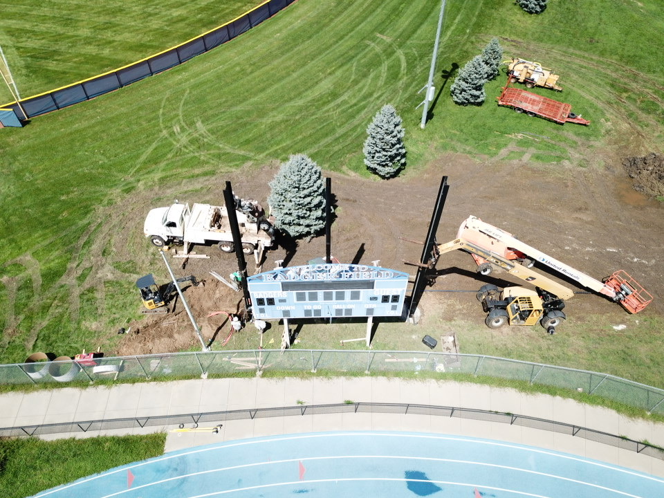 Aerial view of construction equipment and a scoreboard being installed on a sports field, with grass and trees in the background.