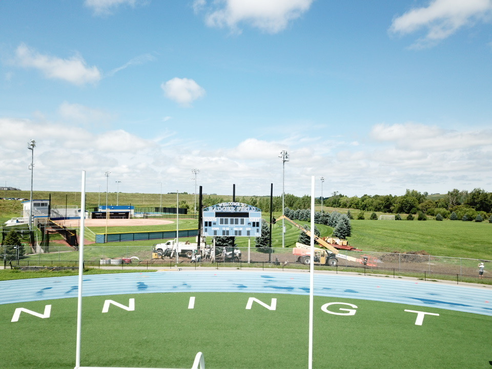 Aerial view of a sports complex featuring a track field, softball field, and scoreboard, with construction equipment and green fields in the background.