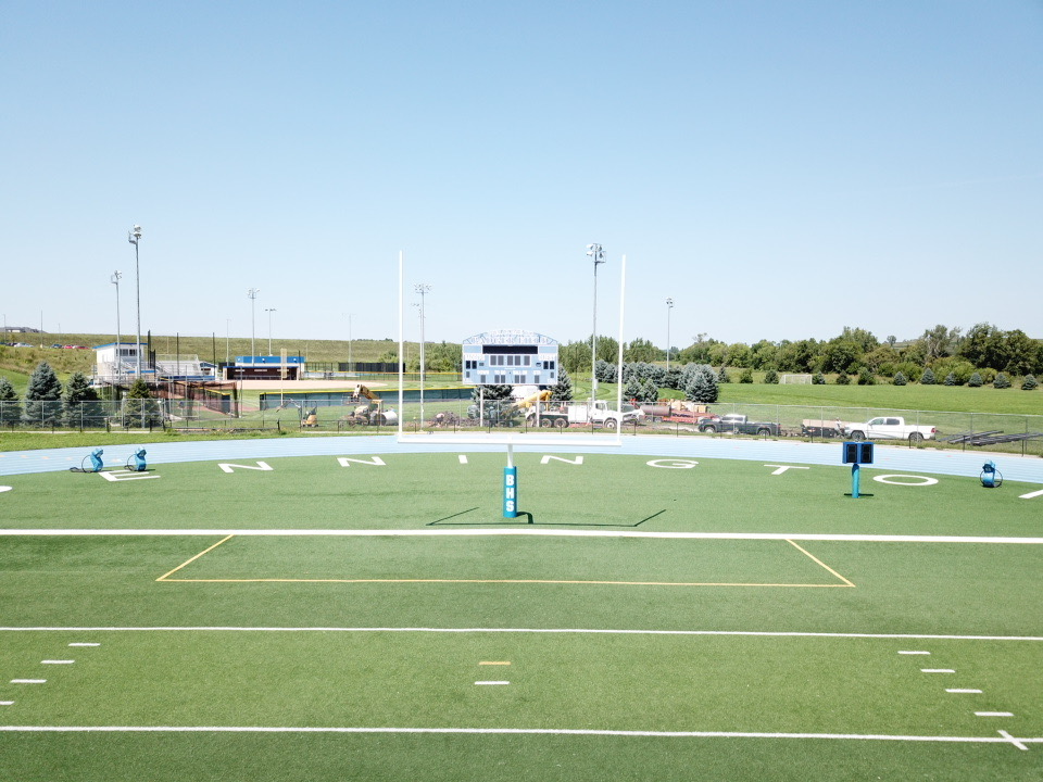 A panoramic view of a football field featuring goalposts and a scoreboard in the background, with construction equipment and vehicles visible beyond the fence.