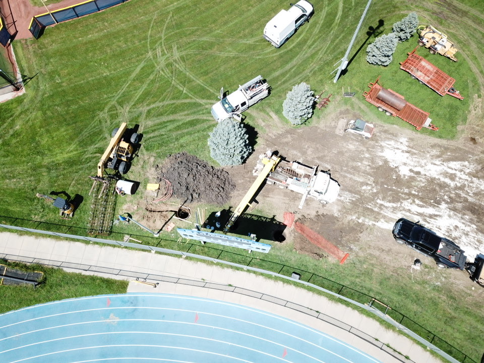 Aerial view of a construction site with heavy machinery, including a digger and a truck, on a grassy field next to a blue running track.