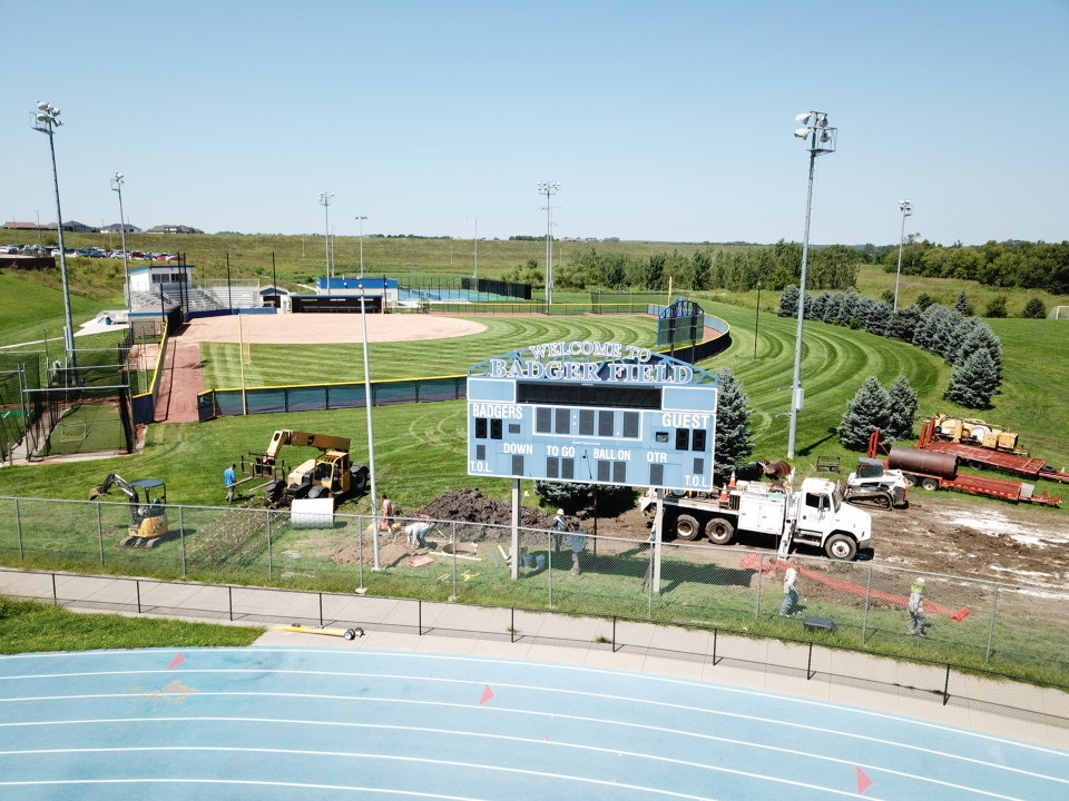 Aerial view of Badger Field featuring a baseball diamond with a scoreboard, nearby construction equipment, and maintenance workers. Lush green grass and trees surround the area.