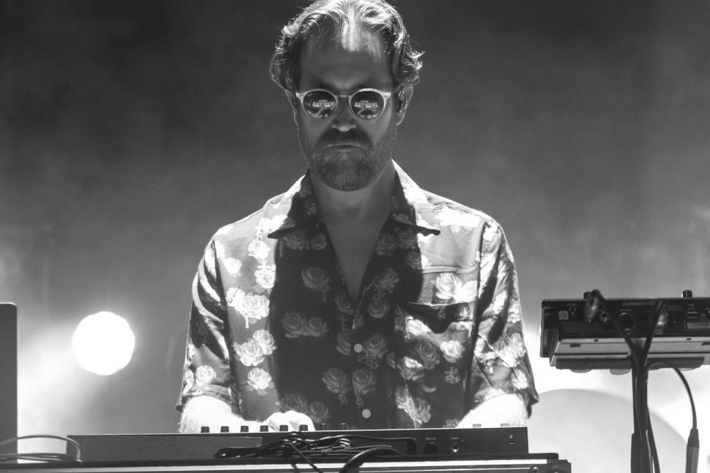 A man wearing sunglasses and a floral shirt, playing a synthesizer on stage with lighting effects in the background.