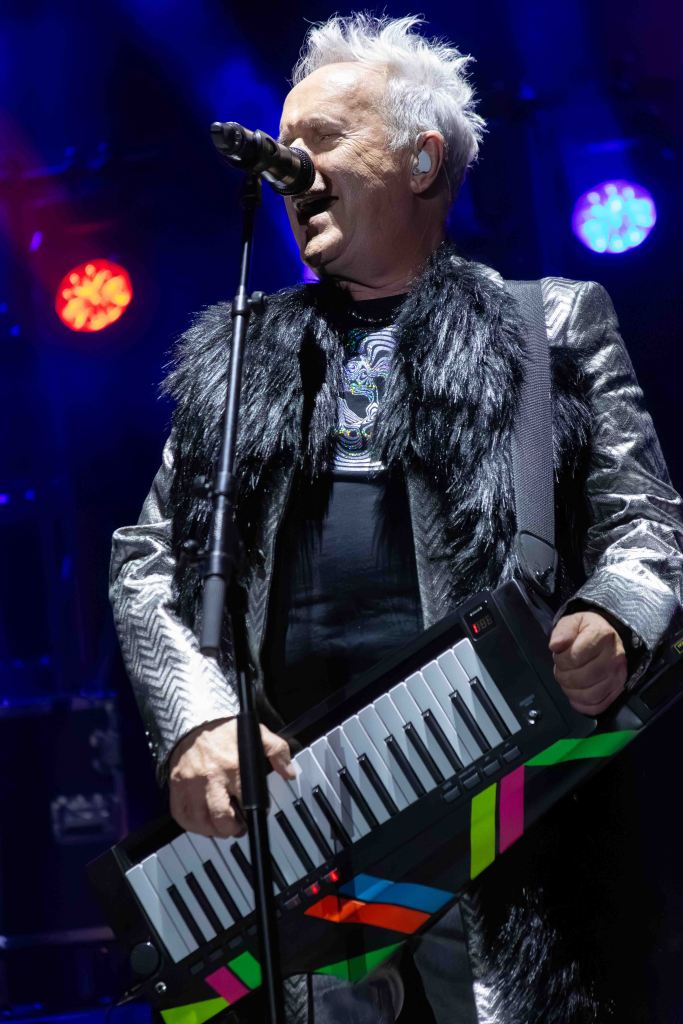 A musician performing on stage, wearing a fur-lined jacket and holding a colorful keytar while singing into a microphone.