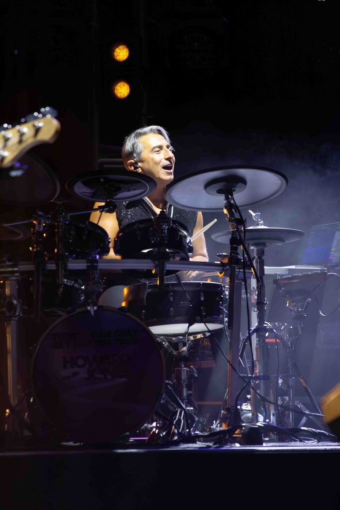 A drummer performing on stage with a lively expression, playing a set of drums under stage lights.