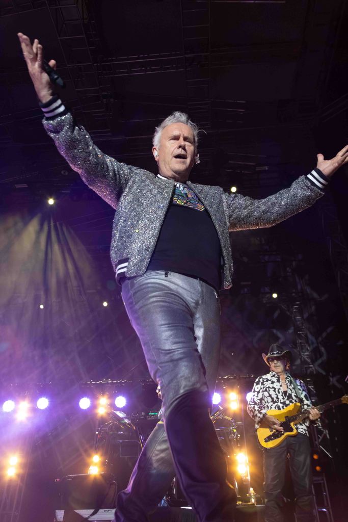 A man in a sparkling silver jacket performs on stage with arms outstretched, while another musician plays guitar in the background.