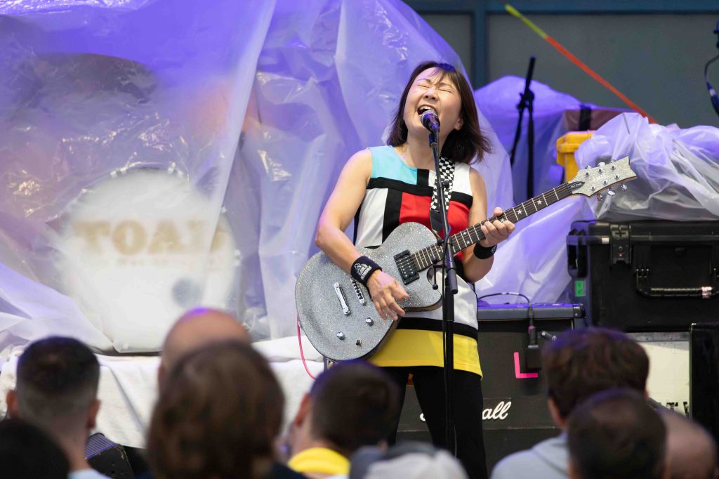 A female musician passionately performing on stage with a guitar, wearing a colorful top, with an audience in front and equipment partially covered in plastic in the background.