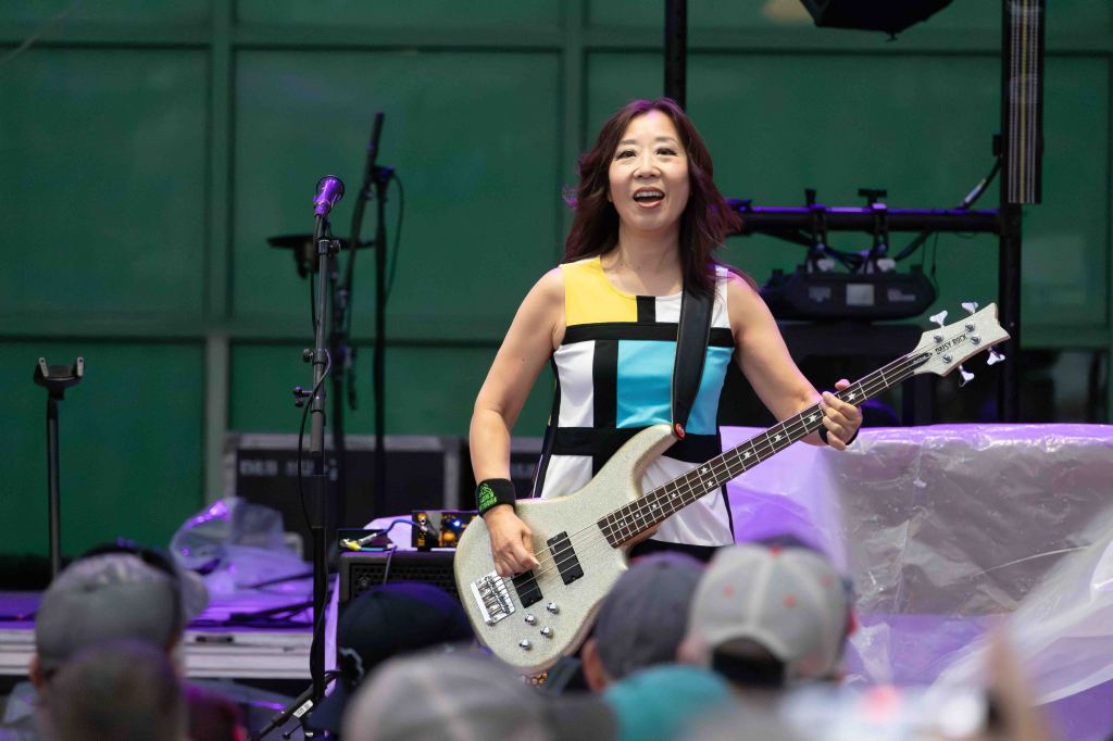 A female musician playing a silver bass guitar on stage, smiling at the audience, wearing a colorful dress with geometric patterns.