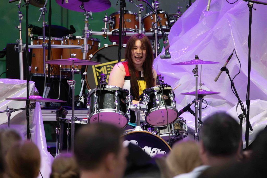 A young female drummer enthusiastically playing on stage, surrounded by various percussion instruments, as an audience watches in the foreground.