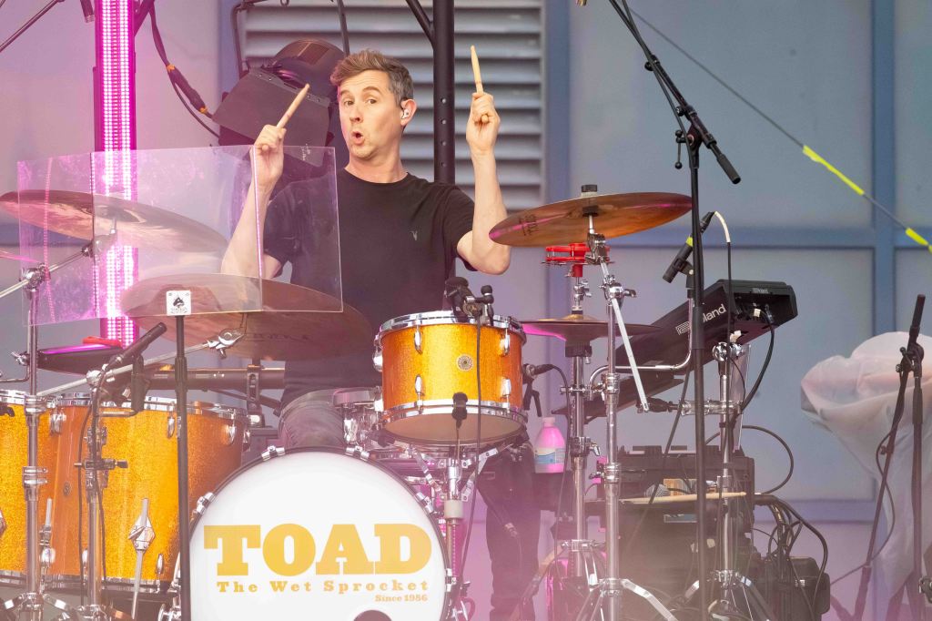 A drummer performing on stage with a clear drum shield, wearing a black shirt and engaging with the audience.